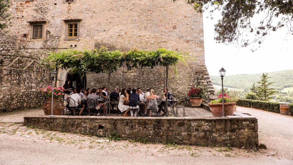 Un gruppo di persone sedute a un tavolo all'aperto, sotto una pergola, in un'antica struttura in pietra circondata da un paesaggio verde.